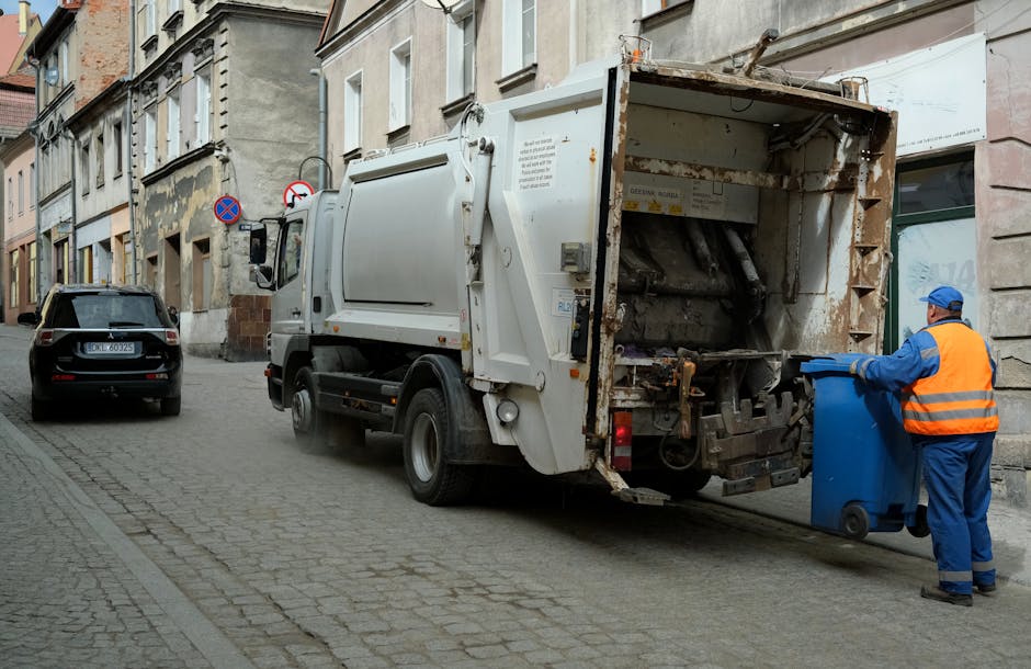 A street scene showing a white waste collection truck operated by Rubbish Removal Kennington, positioned alongside a narrow cobbled road lined with old residential buildings. The truck's rear hatch is open, revealing the mechanical and hydraulic components used for waste compacting. A worker dressed in a blue uniform with an orange high-visibility vest and a blue cap is seen loading a medium-sized blue wheelie bin into the truck. The bin is made of durable plastic with a textured surface and is positioned close to the worker, who is carefully handling it. Next to the truck, a black car is parked on the cobblestone pavement, with its rear end visible. The surrounding buildings have aged, textured facades in shades of beige and grey, with some areas showing signs of wear and patches of weathering. The environment appears to be in an urban area with natural daylight illuminating the scene, emphasizing the practical nature of the waste collection process, which often involves private on-site disposal or alternative rubbish handling methods managed by local waste removal services like Rubbish Removal Kennington.