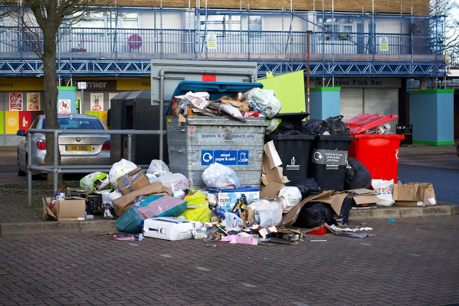 An outdoor scene of overflowing rubbish bins and scattered waste on a paved urban street, with various materials such as cardboard boxes, plastic bags, paper, and packaging spilling onto the pavement around them. The central large grey bin with a blue lid is filled to capacity, with debris piled above its rim, including flattened cardboard, crumpled paper, and plastic wrapping. To its right, there are black and red bins, some overflowing with rubbish, while smaller cardboard boxes and plastic bags are strewn across the ground. In the background, there are storefronts with closed roller shutters, signage indicating shops and services, and a building with blue scaffolding and protective netting suggesting ongoing maintenance or renovation. A silver car is parked behind a metal barrier on the left side of the image, and a tree trunk is visible in the mid-ground. The scene appears to depict a missed or delayed rubbish collection, aligning with guidelines for alternative waste handling and private disposal, as managed by rubbish removal services like Rubbish Removal Kennington. The lighting suggests daytime with natural light illuminating the scene, providing a clear view of the mess and surrounding environment.