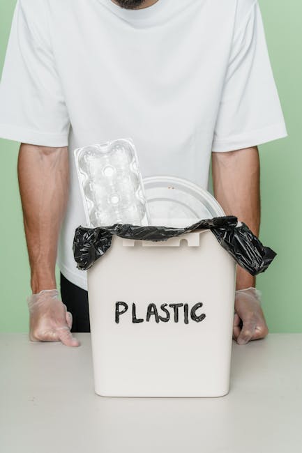 A person wearing a plain white T-shirt and transparent disposable gloves is holding a white plastic container with the word 'PLASTIC' written on it in black marker. The container is topped with a black plastic bag that is tied around the rim. Inside the container, there is a clear plastic packaging tray, resembling a food or egg carton, partially visible above the black bag. The scene is set against a light green background, suggesting an environment focused on waste management or recycling. This setup illustrates the handling and separation of plastic waste, relevant to private rubbish collection and alternative disposal services provided by companies such as Rubbish Removal Kennington. The image emphasizes proper waste sorting practices through the clear and detailed depiction of recyclable plastics in a dedicated container, supporting responsible rubbish removal and environmentally conscious waste handling.