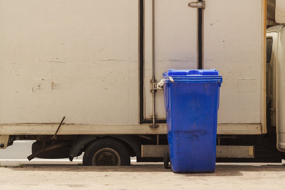 A large blue wheeled rubbish bin made of hard plastic with a textured surface and a slightly darker smudge near the bottom, positioned on the sidewalk adjacent to a white delivery vehicle. The vehicle has a plain, slightly weathered exterior with a small, rusted metal latch and door handle visible on the side. The bin is placed close to a metal pipe running vertically along the vehicle’s side, which is secured with brackets. The scene appears to be outdoors during daytime, with indirect natural lighting illuminating the area, and the background showing a concrete ground with minor cracks. The setting suggests a scene of waste collection or disposal, characteristic of private waste handling services such as those operated by Rubbish Removal Kennington, supporting the practice of independent rubbish collection and on-site clearance for efficient rubbish removal services.