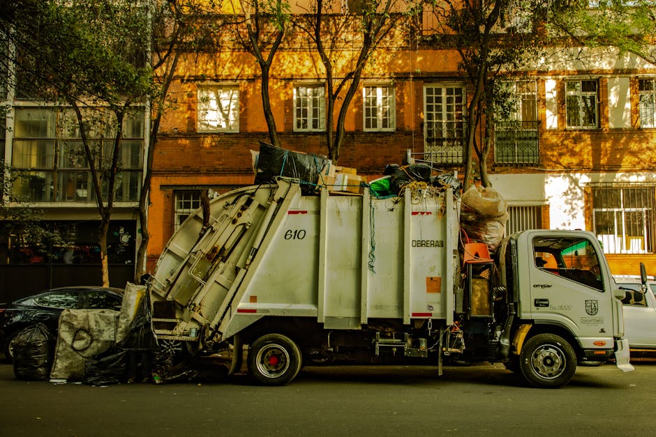 A white rubbish collection truck operated by Rubbish Removal Kennington is parked on a city street in front of a multi-storey brick building with large windows. The truck is loaded with various waste materials, including black trash bags, cardboard boxes, and other debris, some of which are visibly spilling over the top. The vehicle's side panels display the number 610 and the branding of Dorebai. On the left, a black trash bag rests on the pavement beside a large, weathered object wrapped in a grey tarp or cloth. The scene is set in daylight, with shadows cast by surrounding trees and the building, indicating late afternoon or early evening. The environment suggests a typical urban setting where private or independent rubbish collection services are used for waste disposal, aligning with non-local authority waste handling activities associated with alternative rubbish removal. Some trees are visible, with leaves and branches partially blocking the upper parts of the building, contributing to a natural, residential feel.