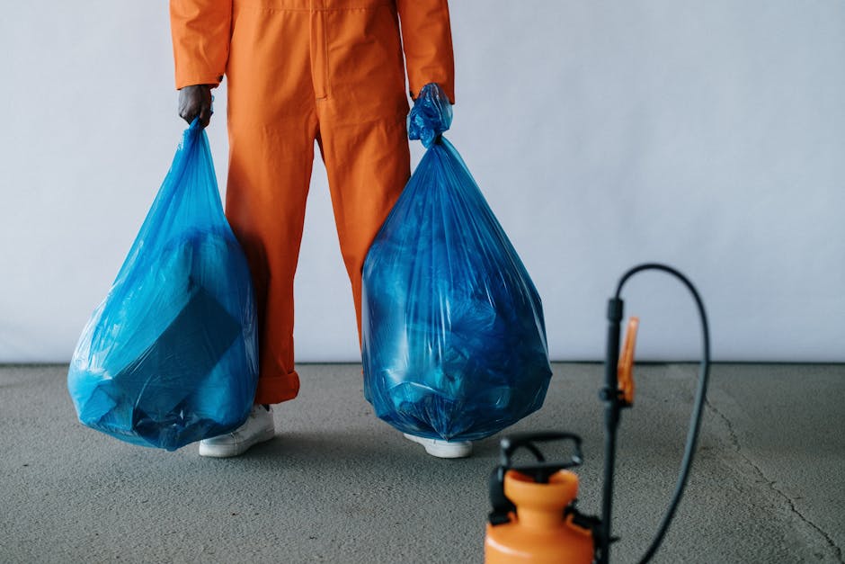 A person wearing orange work trousers and white shoes is standing on a grey concrete surface while holding two large blue plastic rubbish bags, one in each hand. The bags are semi-transparent, revealing a mixture of waste inside, and are tied at the top. The background is plain and neutral, with no distinct features visible. The scene suggests an on-site waste clearance activity, possibly relating to private rubbish collection services such as those provided by Rubbish Removal Kennington as part of their alternative waste handling solutions. The focus on the individual’s lower body and the rubbish bags highlights the physical aspect of rubbish removal, emphasizing the need for proper waste management and disposal, which aligns with the company's services.