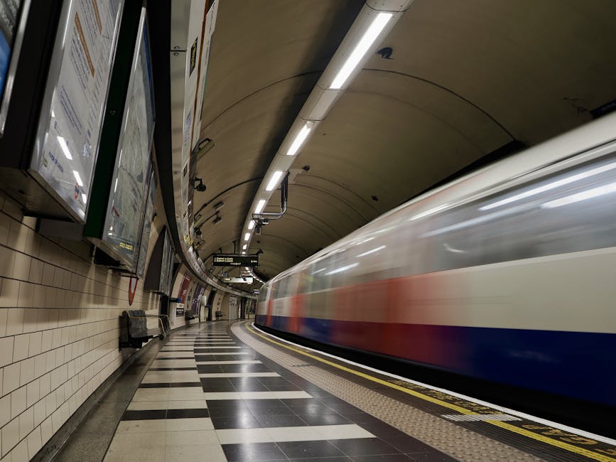 Inside an underground train station with a curved platform featuring tiled walls and black-and-white checkered flooring, a moving train with a sleek, elongated body in white with blue and red horizontal stripes is arriving or departing. The station ceiling is arched and lined with long, white fluorescent lighting fixtures, casting bright illumination across the scene. On the left side, there are electronic display screens and advertisement panels mounted on the wall, with some benches positioned along the platform for waiting passengers. The environment appears clean and modern, with a neutral colour palette predominantly composed of beige, black, white, and metallic tones. This setting exemplifies an underground transit environment, which can be relevant to discussions about alternative waste handling options such as cleaning, maintenance, or designated areas within public infrastructure, relevant to the work of Rubbish Removal Kennington in contexts involving the clean-up or clearance of waste and debris in public spaces.