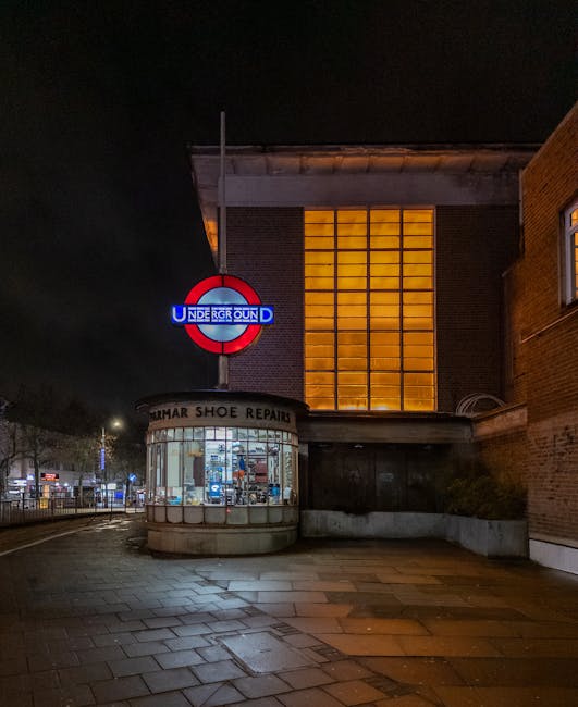 A nighttime exterior view of a building featuring a prominent London Underground roundel sign illuminated in red, white, and blue, positioned above a small, rounded shop front with large glass windows displaying various repair tools and footwear. The shop, identified as 'TUMAR SHOE REPAIR,' is situated at street level and has a brick façade. To the right, a large rectangular window with a warm, amber glow is visible, divided into smaller panes by grid-like frames, contrasting with the surrounding dark brickwork. The pavement in front is wet, reflecting the subtle street lighting, and the background reveals a quiet street scene with distant streetlights and parked vehicles under a clear night sky. The scene suggests a calm urban environment, with the building potentially involved in private or alternative waste handling, such as collection or clearance, relevant to rubbish removal services like those provided by Rubbish Removal Kennington.