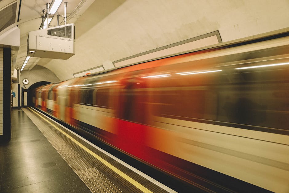 Inside an underground train station with a curved platform featuring tiled walls and black-and-white checkered flooring, a moving train with a sleek, elongated body in white with blue and red horizontal stripes is arriving or departing. The station ceiling is arched and lined with long, white fluorescent lighting fixtures, casting bright illumination across the scene. On the left side, there are electronic display screens and advertisement panels mounted on the wall, with some benches positioned along the platform for waiting passengers. The environment appears clean and modern, with a neutral colour palette predominantly composed of beige, black, white, and metallic tones. This setting exemplifies an underground transit environment, which can be relevant to discussions about alternative waste handling options such as cleaning, maintenance, or designated areas within public infrastructure, relevant to the work of Rubbish Removal Kennington in contexts involving the clean-up or clearance of waste and debris in public spaces.