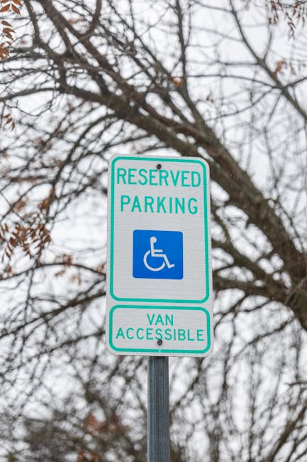 A rectangular signage post positioned outdoors on a metal pole displays a reserved parking sign with a green border, indicating it is designated for disabled parking and accessible via van. The sign features a blue and white wheelchair symbol and the text 'RESERVED PARKING' above it, with the additional note 'VAN ACCESSIBLE' below. The background includes leafless tree branches against a cloudy sky, and the overall scene suggests an urban or residential setting. This type of signage relates to alternative waste handling by highlighting designated spaces that may support access for waste collection vehicles or services like those provided by Rubbish Removal Kennington, ensuring compliance with local regulations and facilitating independent waste disposal.