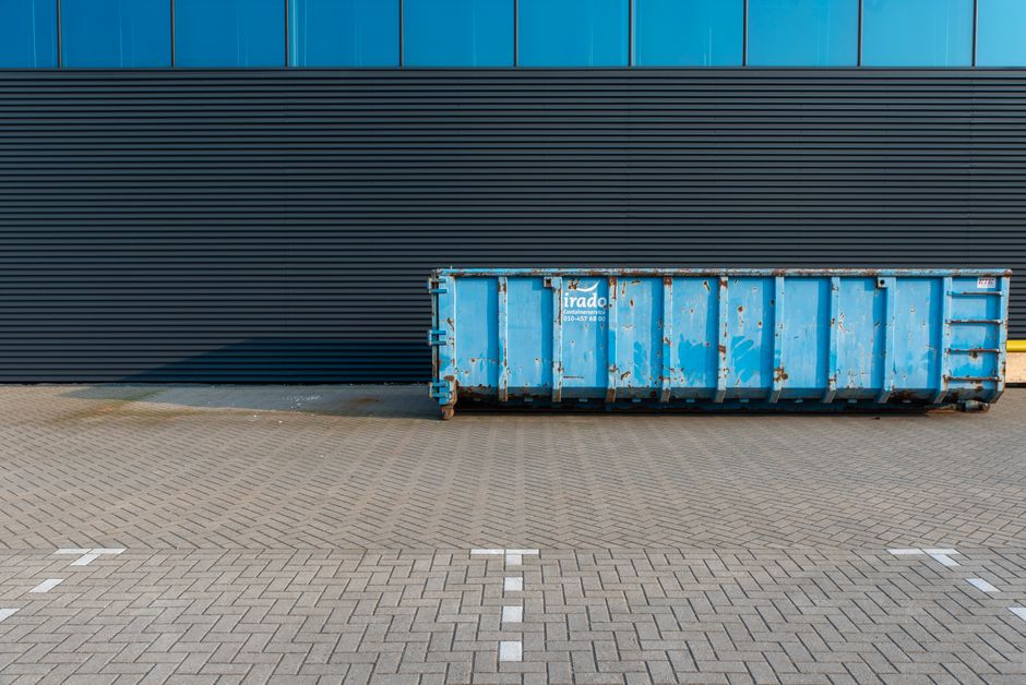 A large blue metal skip bin with a weathered and slightly rusted surface, positioned on a paved concrete surface in front of a modern building with horizontal black and blue siding. The skip is placed close to the building's wall, casting a shadow on the ground, and features visible latches and handling points. The background consists of a sleek, dark-colored wall with horizontal paneling, and the surrounding area includes a section of pavement marked with white lines, indicating designated parking or loading zones. The environment appears clean and industrial, suggesting a setting where waste is temporarily stored for collection or transfer, with Rubbish Removal Kennington providing a controlled and organized waste disposal solution, suitable for alternative on-site clearance or private waste handling services. The lighting is natural, with sunlight casting shadows to the left, emphasizing the textures of the rusty metal and the neat, patterned pavement.
