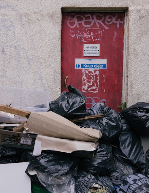 A collection of black plastic rubbish bags and assorted discarded packaging, piled in front of a red metal door set into a beige, rough-textured exterior wall. The door has graffiti tags in white and pink spray paint, as well as a white sign reading 'No Money' and a blue and white 'Keep Clear' sign. A small, green plant is visible growing at the base of the wall near the bottom right corner. The scene appears to be an outdoor area adjacent to a building, potentially a back alley or service entrance, where waste has accumulated for collection or removal. This setting illustrates typical private waste disposal activity in an urban environment, possibly managed by a local rubbish removal service such as Rubbish Removal Kennington, emphasizing the importance of proper rubbish handling and clearance in commercial or residential precincts.