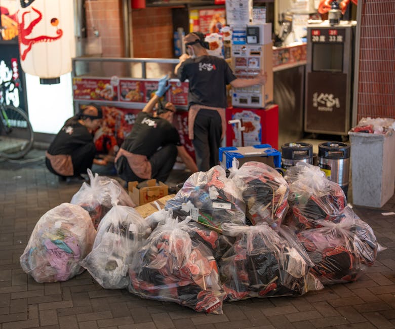 A large pile of clear plastic rubbish bags filled with assorted waste materials, including clothing and packaging, is situated on a paved sidewalk in front of a storefront. The bags are loosely gathered and vary in size, with some showing signs of being partially opened or torn, revealing their contents. Behind the bags, three workers in black uniforms with logo prints on their backs are engaged in loading or organizing additional waste near a red and white food or retail kiosk. The scene is outdoors, illuminated by artificial lighting, and features nearby bins and containers for waste disposal, as well as a small section of a traditional Japanese lantern hanging nearby. The image suggests an environment where private rubbish collection or on-site clearance might be taking place, with the workers handling waste independently outside a commercial premise. The exterior setting includes a tiled pavement and a brick or tile wall backdrop, consistent with urban rubbish removal operations managed by a service like Rubbish Removal Kennington.