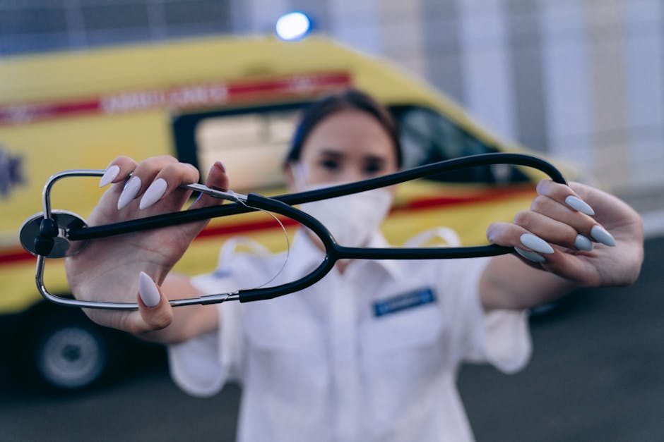A female medical professional wearing a white uniform and a face mask is holding a black stethoscope with silver accents, extending it towards the camera with both hands. Her well-manicured fingernails are painted in a light, neutral color. The background features a yellow emergency vehicle, likely an ambulance, parked outdoors on a paved surface, with its rear side visible and slightly out of focus. The scene is illuminated by natural daylight, creating a clear, professional atmosphere that emphasizes healthcare or emergency services, with the stethoscope symbolizing medical aid. The image subtly relates to the context of independent or private healthcare services, aligning with the themes of urgent care and emergency response, which may connect to the broader topic of flexible, on-site health-related solutions in contrast to public healthcare options.