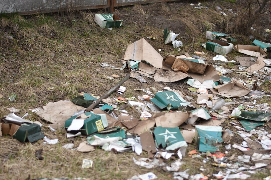 An outdoor scene showing a patch of uneven ground with mixed litter and waste. Scattered around are flattened and torn cardboard boxes, some with printed branding and logos, along with a few plastic bottles and small pieces of paper. The cardboard boxes are mainly in shades of beige and green, with a rough, folded texture. Some boxes have creases and dents, indicating they have been discarded after use. The ground features patchy grass and bare soil with a slight incline, and in the background, there is a wooden or metal fence partially visible. The overall environment appears neglected, with rubbish strewn randomly across the area, typical of illegal dumping or fly-tipping. This scene illustrates a typical example of waste that might require professional rubbish removal services, such as those provided by Rubbish Removal Kennington, especially for private disposal or alternative waste handling outside official council collections.