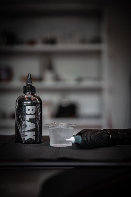 A close-up image of a black plastic squeeze bottle with the word 'BLACK' printed vertically in bold white letters, positioned on a dark surface. Next to the bottle is a small transparent plastic container filled with a clear liquid and a tattoo machine with a textured black grip lying horizontally, with its needle tip directed towards the liquid container. The background features a blurred interior space with indistinct shelving and objects, suggesting an environment like a tattoo studio or workspace. The scene is lit with soft, diffuse lighting, emphasizing the textures of the bottle's glossy finish and the tattoo machine's grip, while the environment remains unobtrusive. The arrangement indicates an area designated for artistic or cosmetic procedures, possibly related to ink or pigment handling, aligning subtly with services like those offered by Rubbish Removal Kennington who handle various waste-related activities, including managing equipment and materials associated with such environments. The overall workmanship reflects attention to detail typical in professional settings concerned with cleanliness and safety, relevant for private or alternative waste handling contexts involving equipment and consumables.